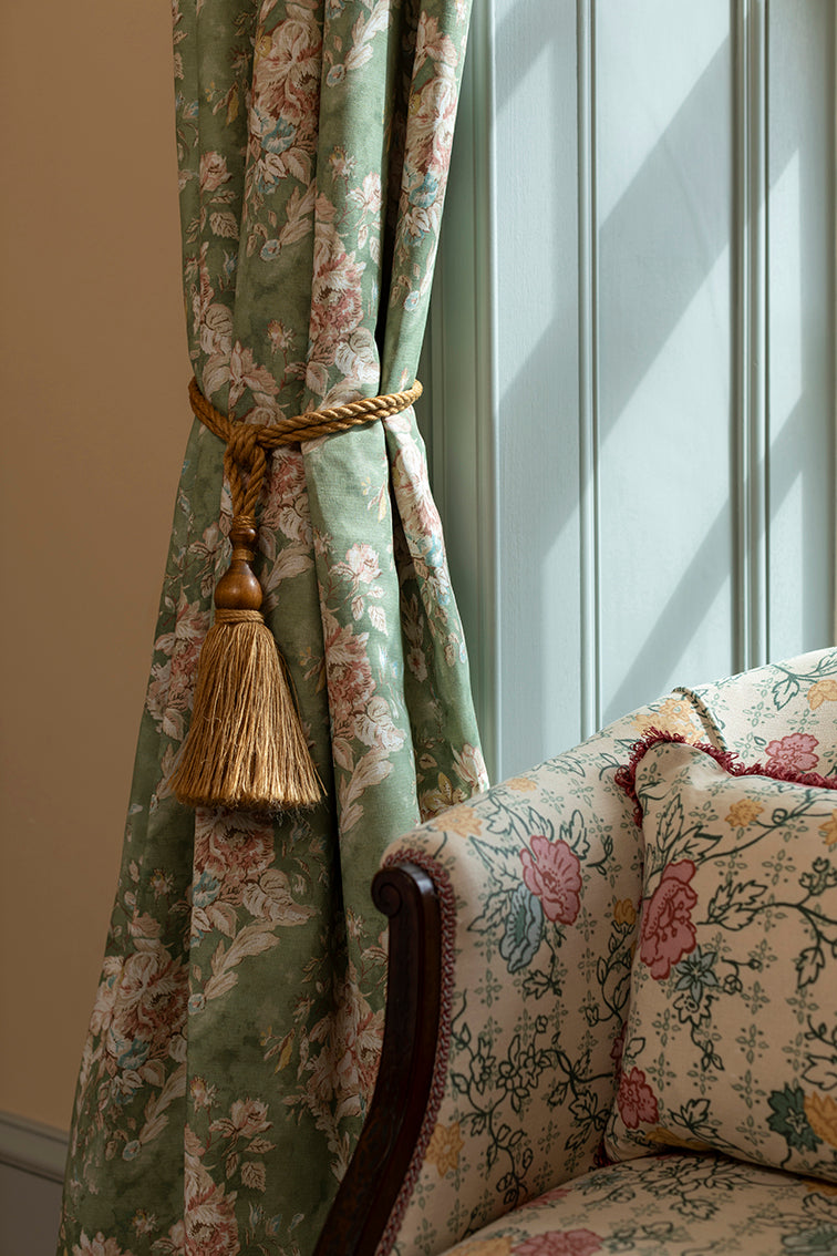 Floral-patterned curtain with a tassel and matching sofa in a room with sunlight filtering through.